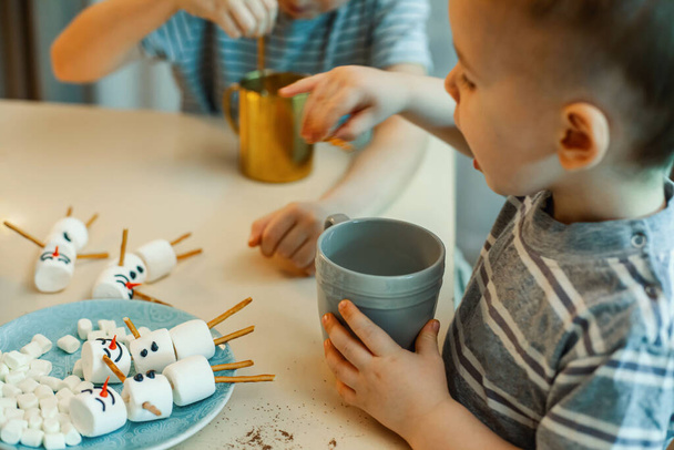 Children make snowmen from marshmallows and salted straws, drink cocoa in the home kitchen. Preparing for Christmas and New Year. - Valokuva, kuva