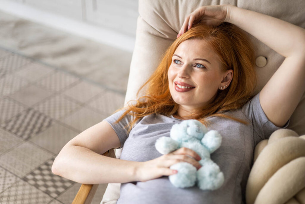 Top view happy young redhead pregnant woman lying on comfortable armchair with cute bear toy - Photo, image