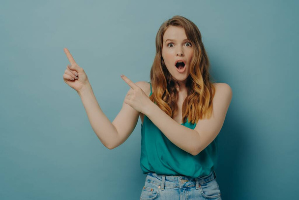 Удивлённая молодая женщина в повседневной одежде, указывающая на правую сторону указательными пальцами, изолированная на голубом - Фото, изображение