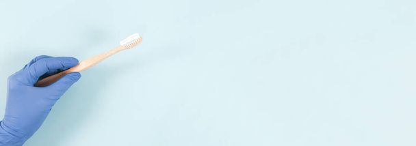 Uma mão enluvada segura uma escova de dentes de bambu com pasta à esquerda contra um fundo azul suave com espaço de cópia à direita, vista lateral close-up. - Foto, Imagem