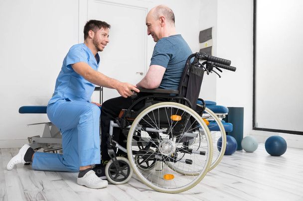 Mannelijke fysiotherapeut helpt een patiënt met een handicap die een rolstoel gebruikt om op te staan in het revalidatieziekenhuis. - Foto, afbeelding
