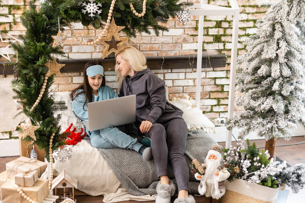 Valkoihoinen nainen tyttärensä kanssa jouluna, jolla on videokeskustelu ystävien ja perheen kanssa kannettavan tietokoneen näytöllä, istuu sohvalla kotona syleillen. sosiaalinen etäisyys joulun kovan 19-pandemian aikana. - Valokuva, kuva