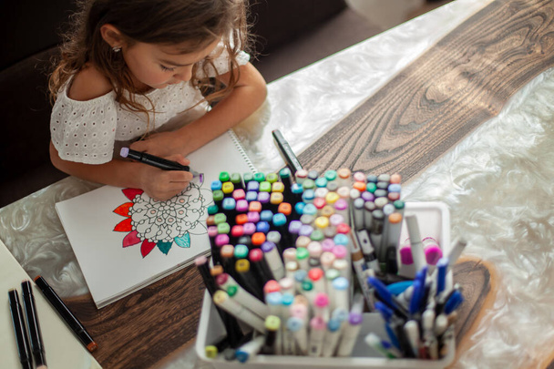 A little cute girl draws a circular mandala pattern in the album with art markers. - Foto, imagen