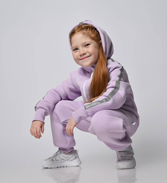 Portrait of cute smiling red-haired kid girl in pink jumpsuit sitting squatted with her hood on her head - Valokuva, kuva