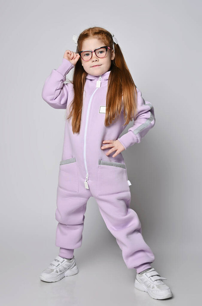 Cool red-haired kid girl in glasses and pink jumpsuit with zipper and big pockets stands with legs wide apart - Фото, изображение