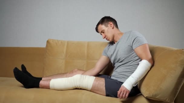 El hombre se sienta en el sofá con la rodilla lesionada y el antebrazo roto - Imágenes, Vídeo