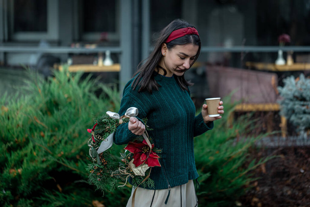 Egy fiatal nő karácsonyi koszorúval és egy csésze kávéval a városi sétányon.. - Fotó, kép