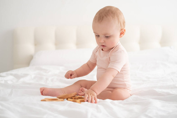 Açık pembe elbiseli bebek beyaz yatakta tahta oyuncaklarla oynuyor. - Fotoğraf, Görsel