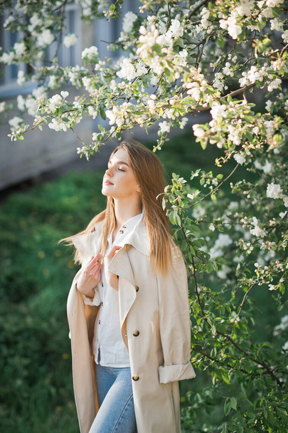 A beautiful girl in a long beige raincoat basks in the sun. - Фото, изображение