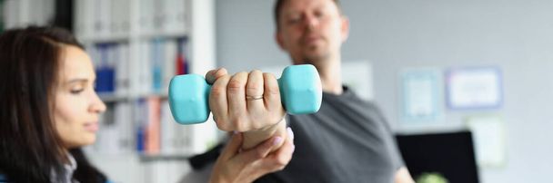 Rehabilitation therapist helping disabled man in wheelchair to lifting dumbbell in closeup - Photo, Image