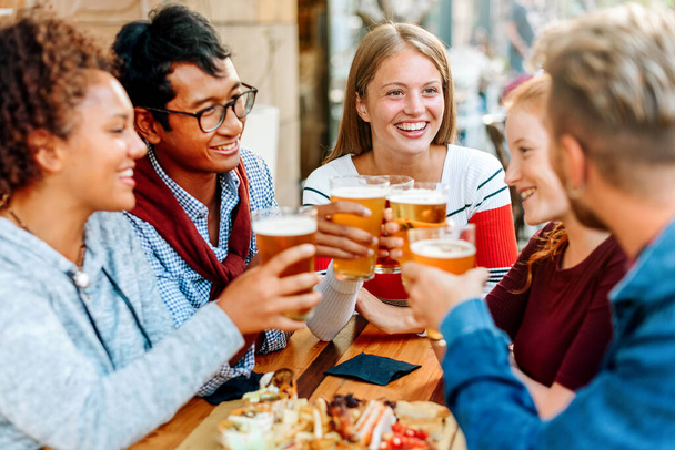 Groupe de jeunes amis divers dégustant une bière ensemble levant leurs verres dans un pain grillé avec une jeune femme blonde souriante - Photo, image