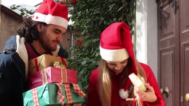 Un joven con sombrero de Papá Noel trae regalos a su novia. Ella sonríe a su novio y lo besa en la mejilla. - Imágenes, Vídeo