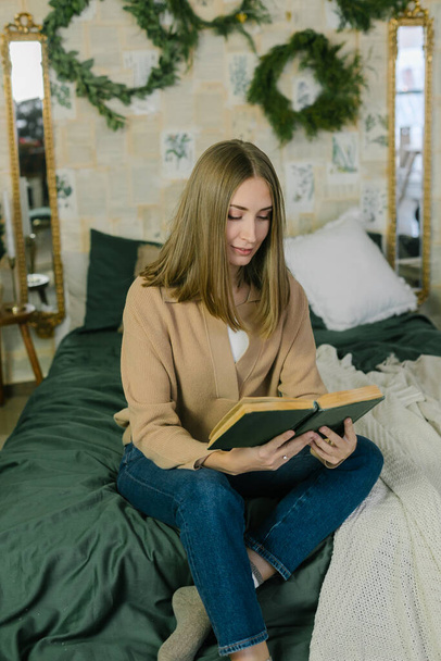Young relaxed caucasian woman in casual outfit reading book while relaxing on bed at home in cozy decorated bedroom at home. Happy Winter Christmas Holidays - 写真・画像