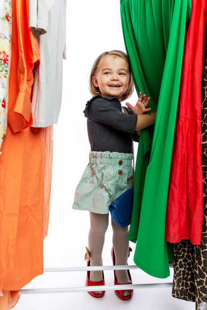 Smiling little girl in red shoes of mom among dresses on hangers. Delight, happiness and fashion. Vertical. - Foto, Imagem