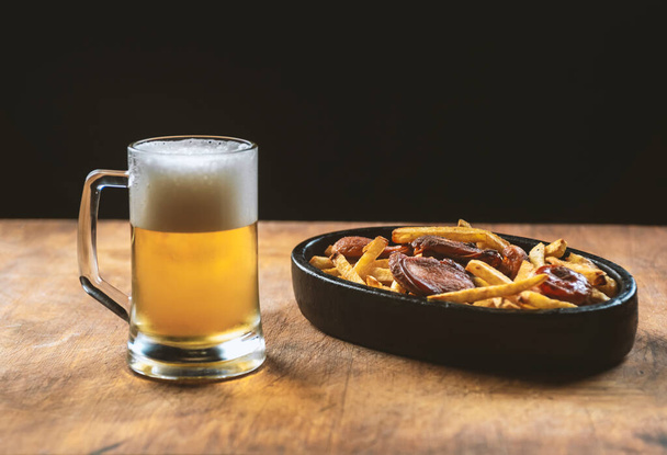 foam in a glass with beer and fried sausages with potatoes on a wooden background.Oktoberfest. - Foto, afbeelding