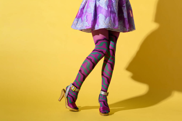 Crop unrecognizable female with sexy legs wearing fashionable purple ornamental collant with mini skirt and high heeled shoes on yellow background with shadow - Fotografie, Obrázek