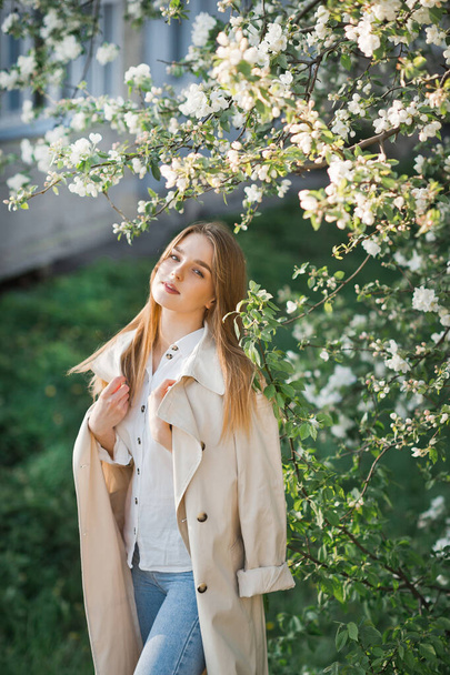 A beautiful girl in a long beige raincoat basks in the sun. - Fotoğraf, Görsel