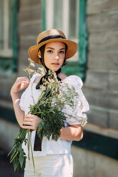 A girl in a hat and a white blouse walks against the background of old wooden houses. - Fotoğraf, Görsel