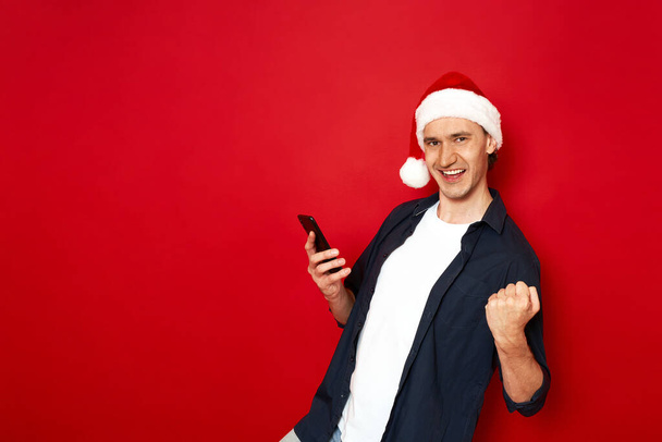 vzrušený, radostný muž ve vánočním klobouku v neformálním oblečení s telefonem v ruce slaví úspěch ukazuje vítězství gesto izolované na červeném pozadí prostoru pro text. koncept - vítězství, technologie, lidé - Fotografie, Obrázek