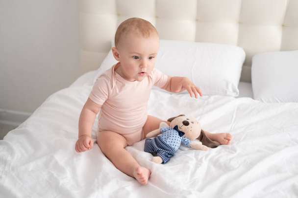 Açık pembe elbiseli bebek beyaz yatakta yumuşak oyuncaklarla oynuyor. - Fotoğraf, Görsel