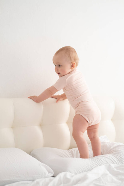 Schattig baby meisje in bodysuit staan houden bed frame, leren lopen op wit beddengoed op bed - Foto, afbeelding