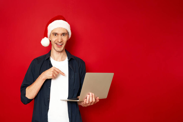 ένας ενθουσιασμένος, χαρούμενος άντρας με χριστουγεννιάτικο καπέλο σε casual ρούχα με φορητό υπολογιστή στο χέρι δείχνει το δάχτυλό του προς τη συσκευή στον κόκκινο τοίχο του στούντιο. concept - τεχνολογίες, άνθρωποι, προσφορές προσφορών Πρωτοχρονιάς - Φωτογραφία, εικόνα