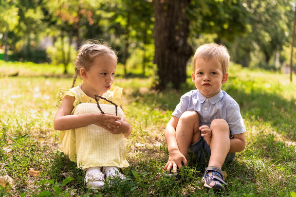 Dwa szczęśliwe uśmiechnięte wesołe maluch przedszkola bliźniaki rodzeństwo dzieci brat siostra chłopiec dziewczyna siedzi razem na trawie trawnik w parku w słoneczną gorącą letnią pogodę. dzieciństwo, przyjaźń, koncepcja rodziny - Zdjęcie, obraz