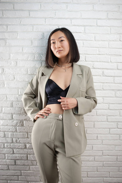 una donna asiatica alla moda in un pantalone elegante posa contro un muro di mattoni bianchi - Foto, immagini