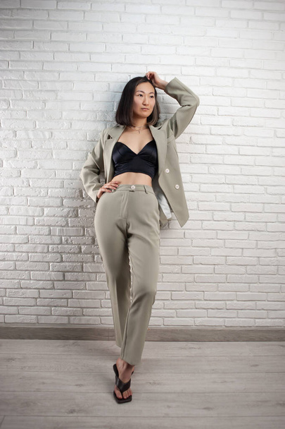 a fashionable Asian woman in a stylish pantsuit poses against a white brick wall - Fotoğraf, Görsel