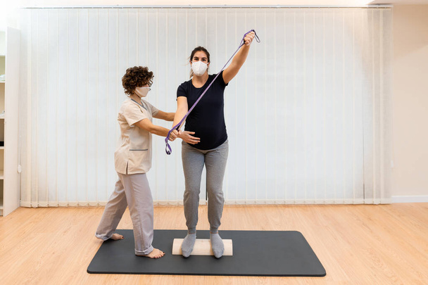 A pregnant woman standing on a proprioceptive trunk or wooden roller with an elastic band with physiotherapist - Fotografie, Obrázek