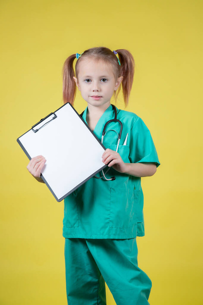 Porträt eines ernsthaften kleinen kaukasischen Mädchens im grünen Arztkittel mit einer Tablette in der Hand, isoliert auf gelb - Foto, Bild