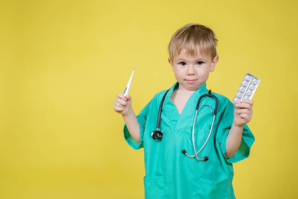 Portrait of little caucasian kid dressed in doctors green coat holds blisters with pills in hand on yellow background - Fotó, kép