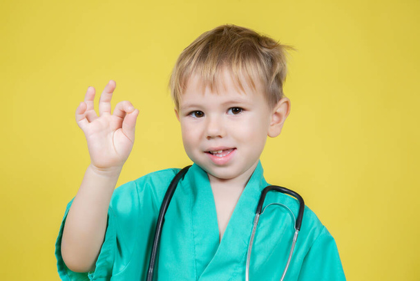 Portrait of cute little caucasian boy dressed in doctors green showing okay ok gesture on yellow background - Foto, imagen