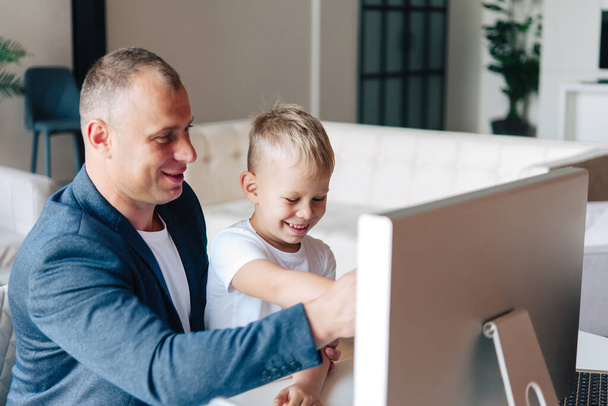 Happy father sits and works at the computer, next to him is a happy preschool child, looking at the monitor and smiling. - Foto, Imagen