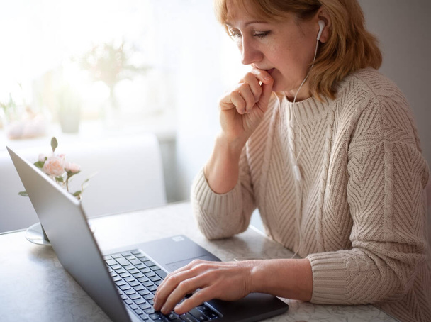 Een Europese vrouw van middelbare leeftijd studeert thuis. Als ze goed naar het laptopscherm kijkt, luistert ze aandachtig naar de informatie op haar koptelefoon. Werkplaats in keuken met laptop. - Foto, afbeelding