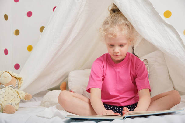 a cute girl with curly hair reads a book in a hut from a sheet on the bed, little girl having fun and playing in her tent, Hut in the kids room - Фото, зображення
