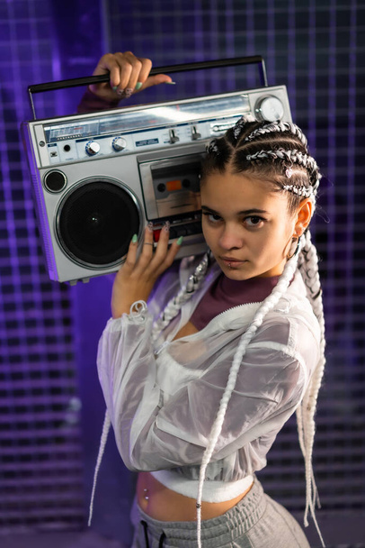 Portrait of a young girl with white braids with a boombox on her shoulders, urban background, purple leds, 80s style, retro photography, vertical photo - Photo, Image