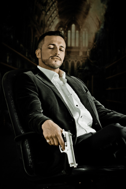 Elegant man sitting in a chair holding  gun over dark background - Fotó, kép