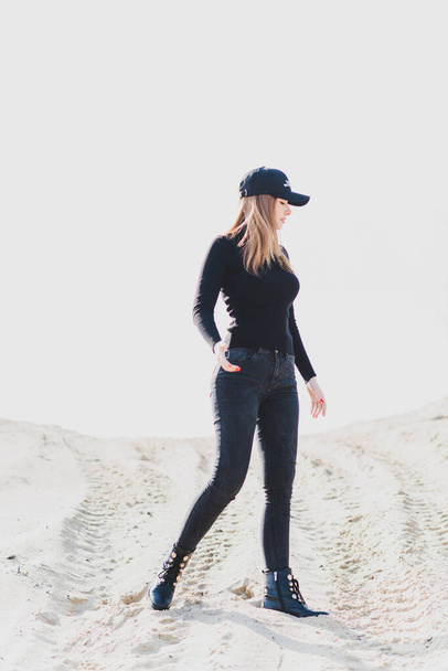Vrouw draagt zwarte coltrui, jeans en honkbalpet, meisje in de zandwoestijn. Modieuze casual stijl, mode en kleding concept van moderne look - Foto, afbeelding