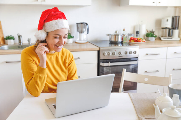 サンタの帽子をかぶっている笑顔の女の子は、ウェブカメラで家族に電話しています。休日に仮想会議チャットを使用して自宅でキッチンに座ってノートパソコンを持つ女性。ハッピークリスマスと新年の新しい通常. - 写真・画像