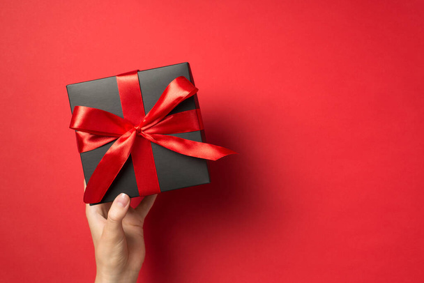 Фото от первого лица с изображением руки, держащей черный подарочный коробок с красной ленточкой на изолированном красном фоне с копирайтом - Фото, изображение