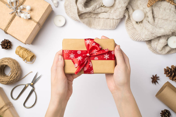 First person top view photo of woman's hands holding craft paper giftbox with red ribbon bow over scarf christmas tree balls and handicraft tools on isolated white background - Photo, Image