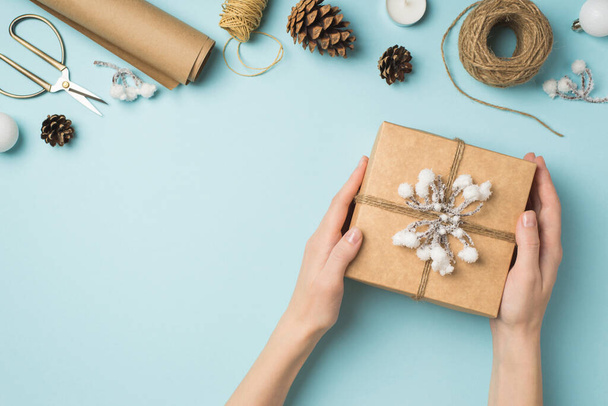 First person top view photo of hands holding kraft paper giftbox decorated with snow branch and twine white christmas tree balls handicraft tools on isolated pastel blue background with empty space - Photo, Image