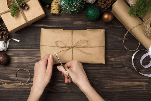 First person top view photo of woman's hands packing kraft paper giftbox with twine bow scissors green gold christmas tree balls pine twigs and handicraft tools on isolated wooden desk background - Fotografie, Obrázek