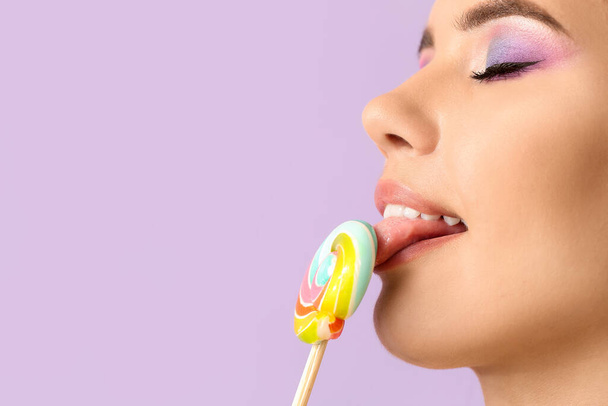 Beautiful young woman eating sweet lollipop on lilac background, closeup - Photo, Image