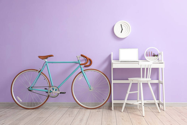 Современное рабочее место с ноутбуком и велосипедом возле лиловой стены - Фото, изображение