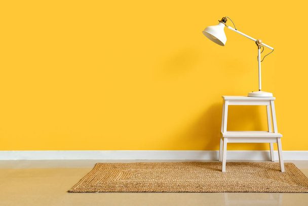 Білий ступінчастий табурет з сучасною лампою біля жовтої стіни
 - Фото, зображення