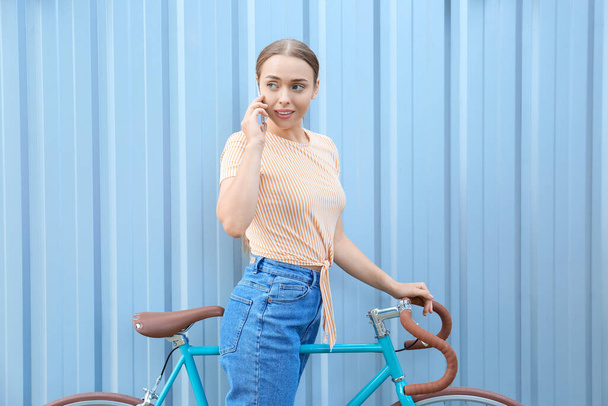 Piękna nastolatka z rowerem rozmawia przez telefon komórkowy w pobliżu niebieskiego ogrodzenia - Zdjęcie, obraz