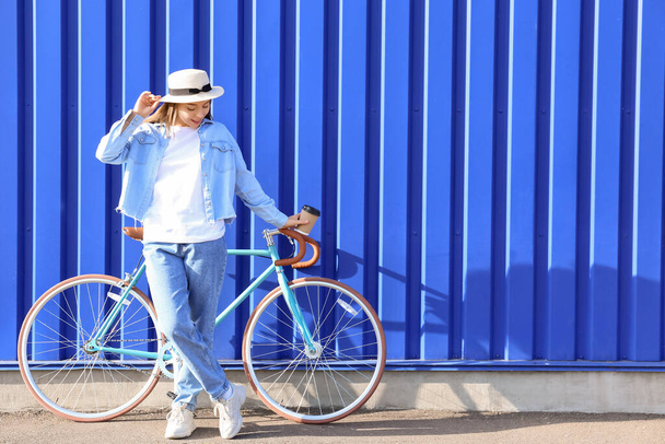 Красивая девочка-подросток с чашкой кофе и велосипедом возле синего забора - Фото, изображение