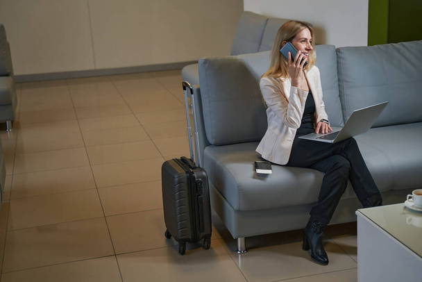 Bilgisayarlı gülümseyen kadın otelde akıllı telefondan konuşuyor. - Fotoğraf, Görsel